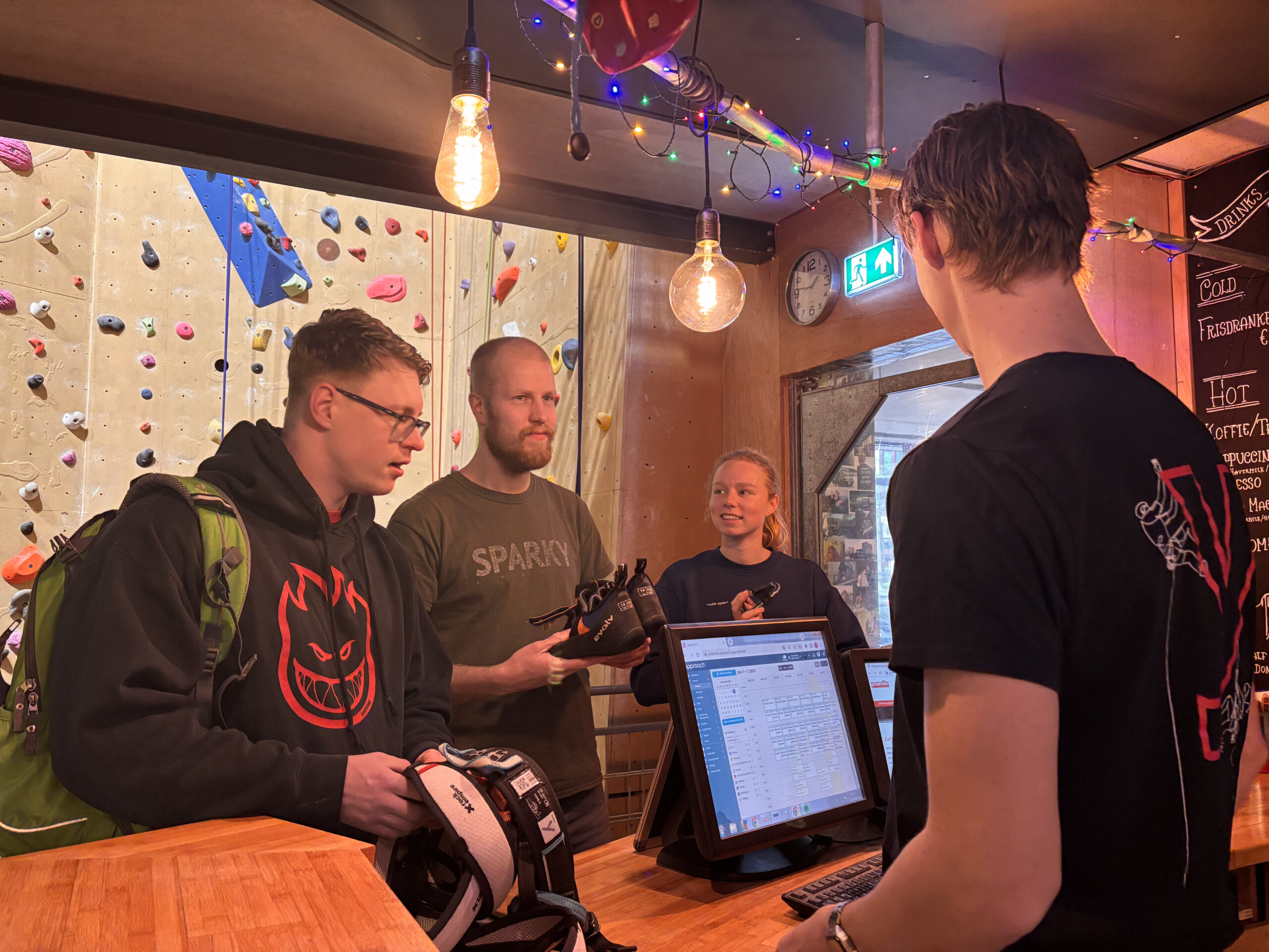 *"Climbers at an indoor gym reception, checking in with staff. A climbing wall with colorful holds is visible in the background."*